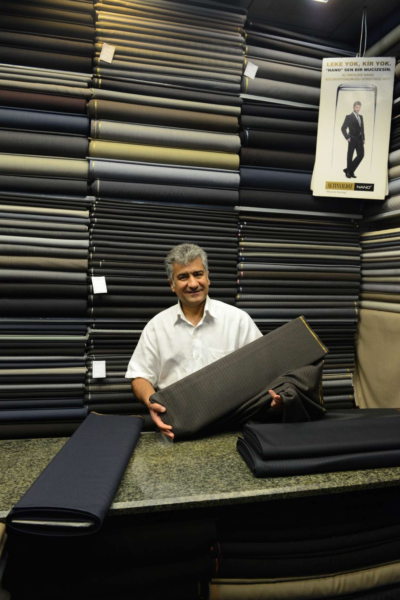 Mohammad Rezai holds fabric in his men's suit shop surrounded by stacked bolts of cloth.