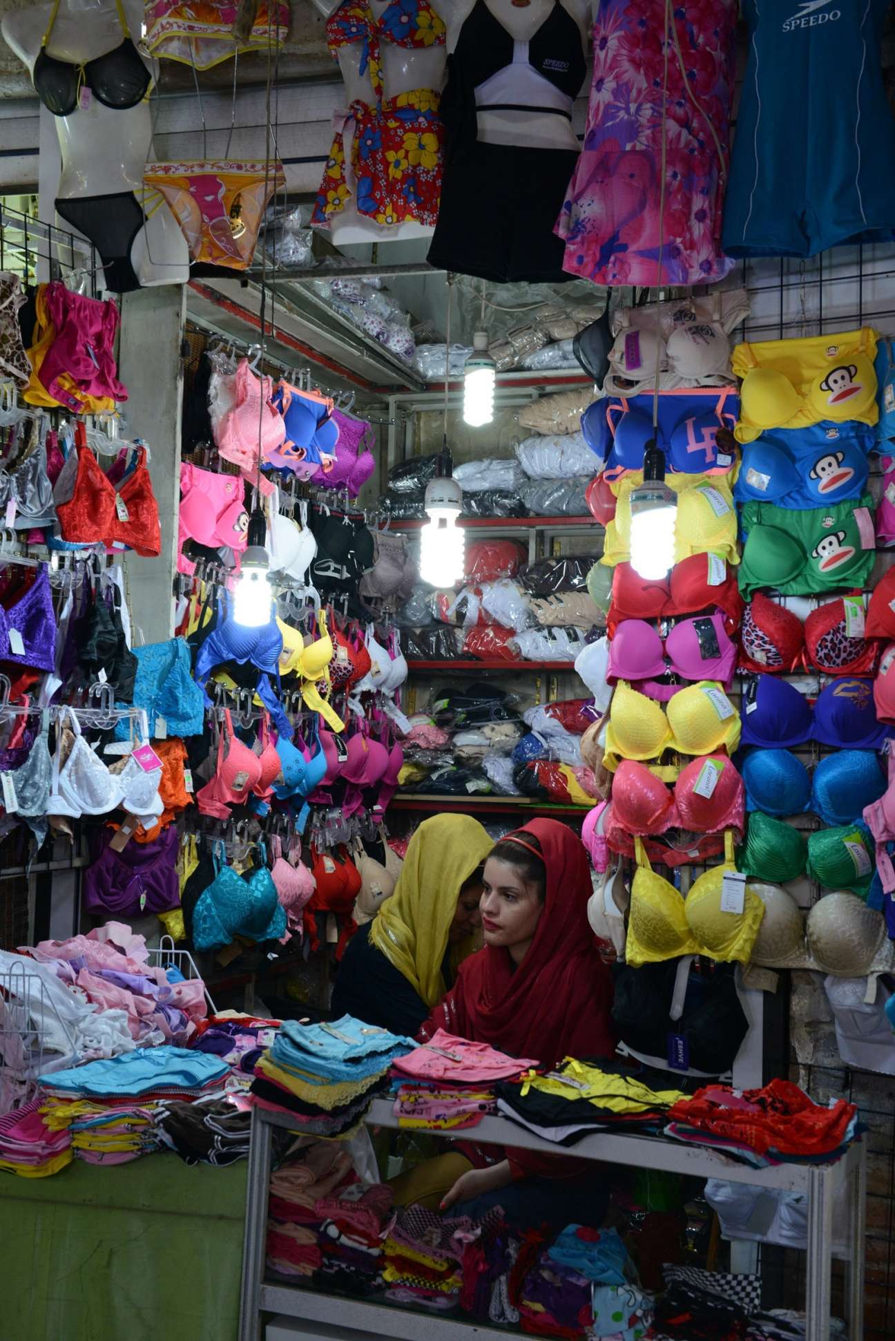 Woman in red headscarf inside a shop stall displaying colorful bras and underwear on walls and shelves.