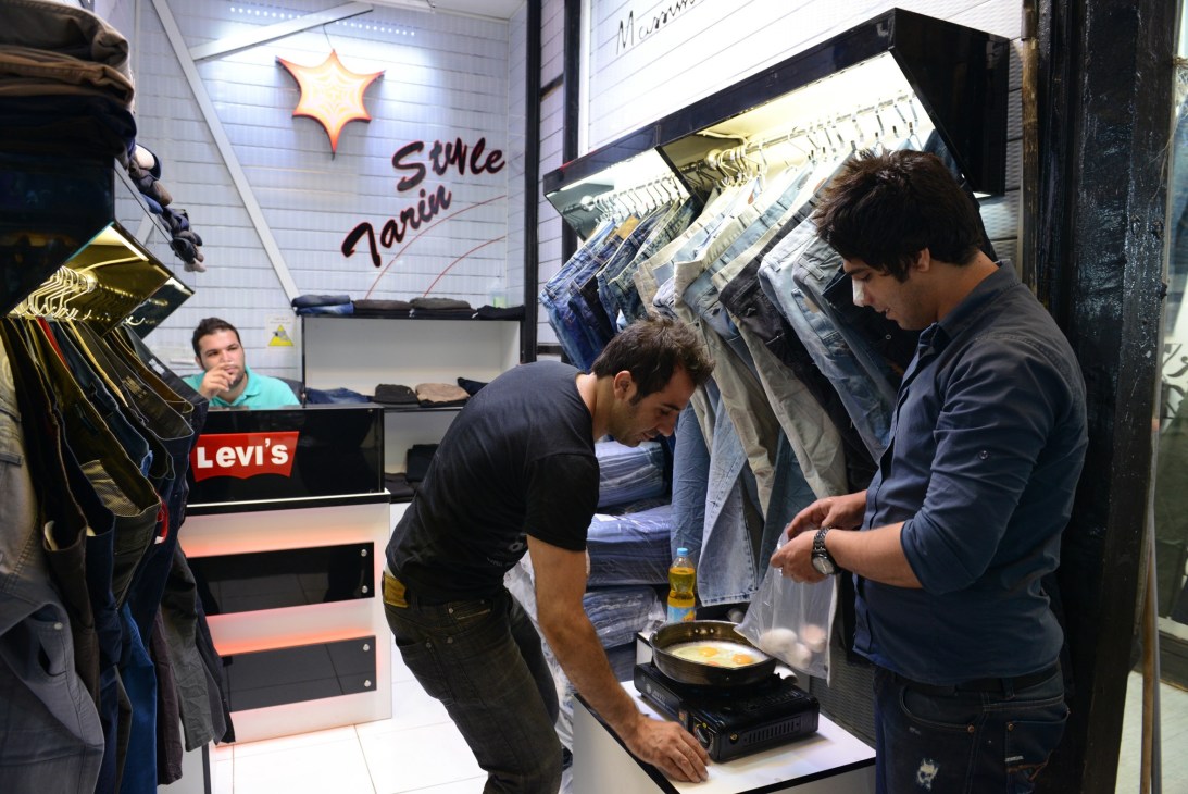 Men shopping at a fake Levi's jeans stall with fried eggs cooking on a portable stove in Tehran's Grand Bazaar.