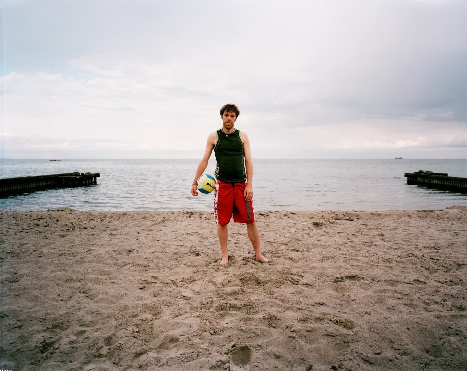 Volleyball player Rasmus Johanssen standing on a beach in Klampenborg holding a volleyball, with the sea and wooden piers visible behind him.