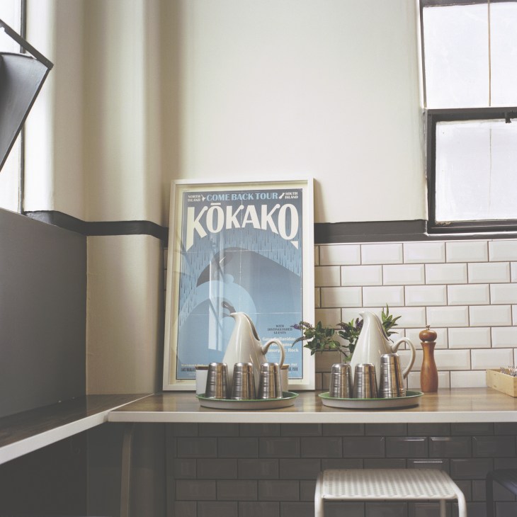 Kokako café interior with metal coffee cups, potted plant, and vintage poster on white subway tile wall.