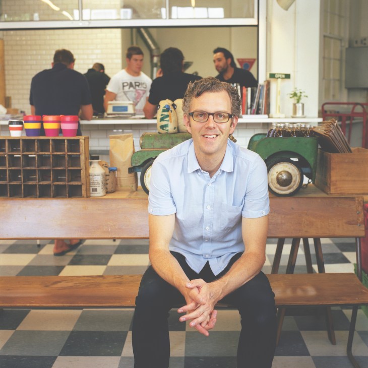 Man in light blue shirt sitting at wooden table in Kokako café with staff and colorful cups visible behind counter.