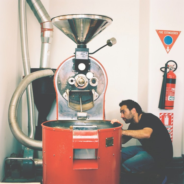 Man operating a red coffee roaster machine with industrial piping and fire extinguisher visible.