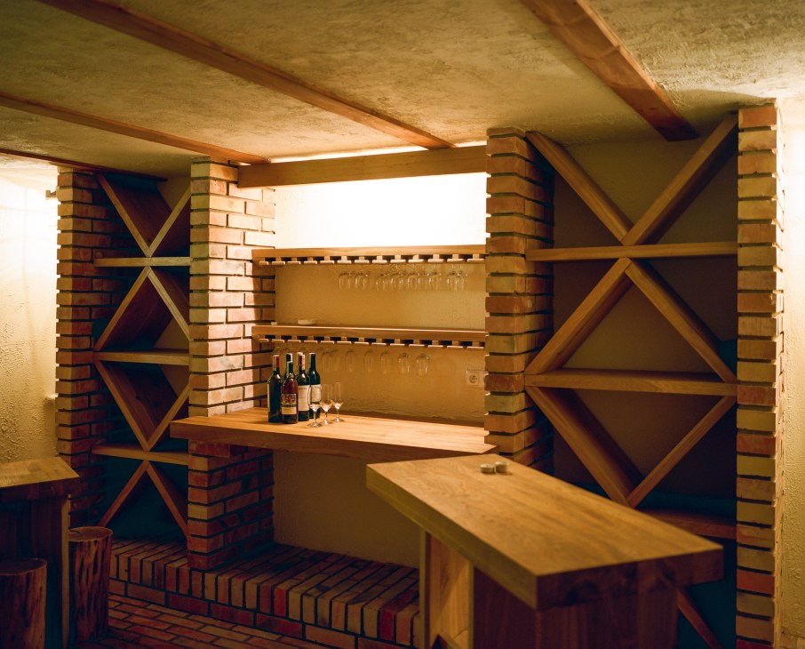 Wine cellar with brick pillars, wooden X-braces, wine bottles and glasses on shelves.