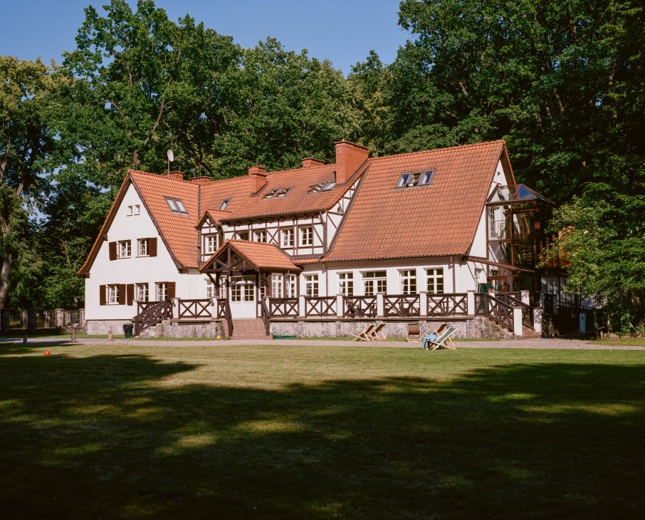 Half-timbered Prussian-style main building with terracotta roof and wooden terrace overlooking manicured grounds and forest backdrop.