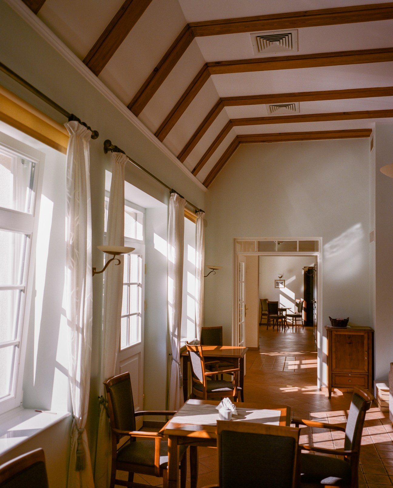 Bright hallway with exposed wooden ceiling beams, white curtains on large windows, wooden furniture, and doorways leading to other rooms.