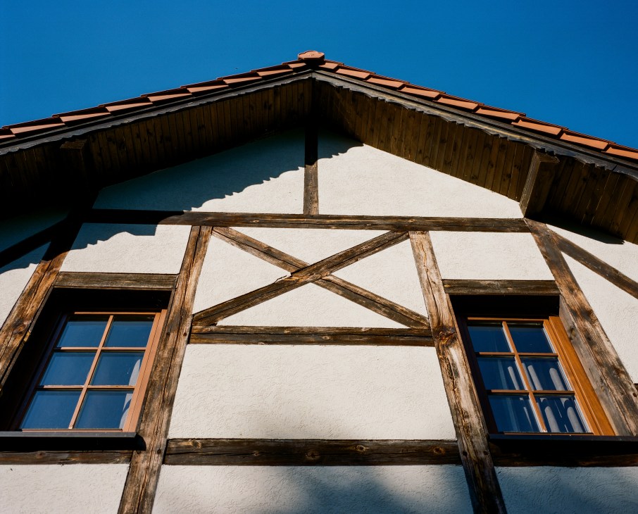 Half-timbered Prussian building facade with white walls, dark wooden beams, terracotta roof tiles, and blue-framed windows against clear sky.