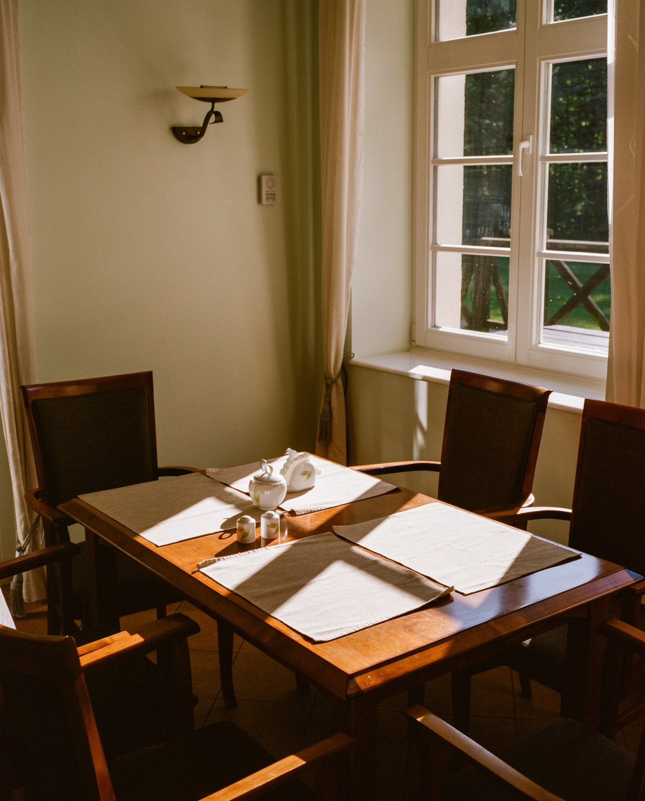 Sunlit dining area with wooden table, dark chairs, and large windows overlooking forest grounds at Nibork retreat.