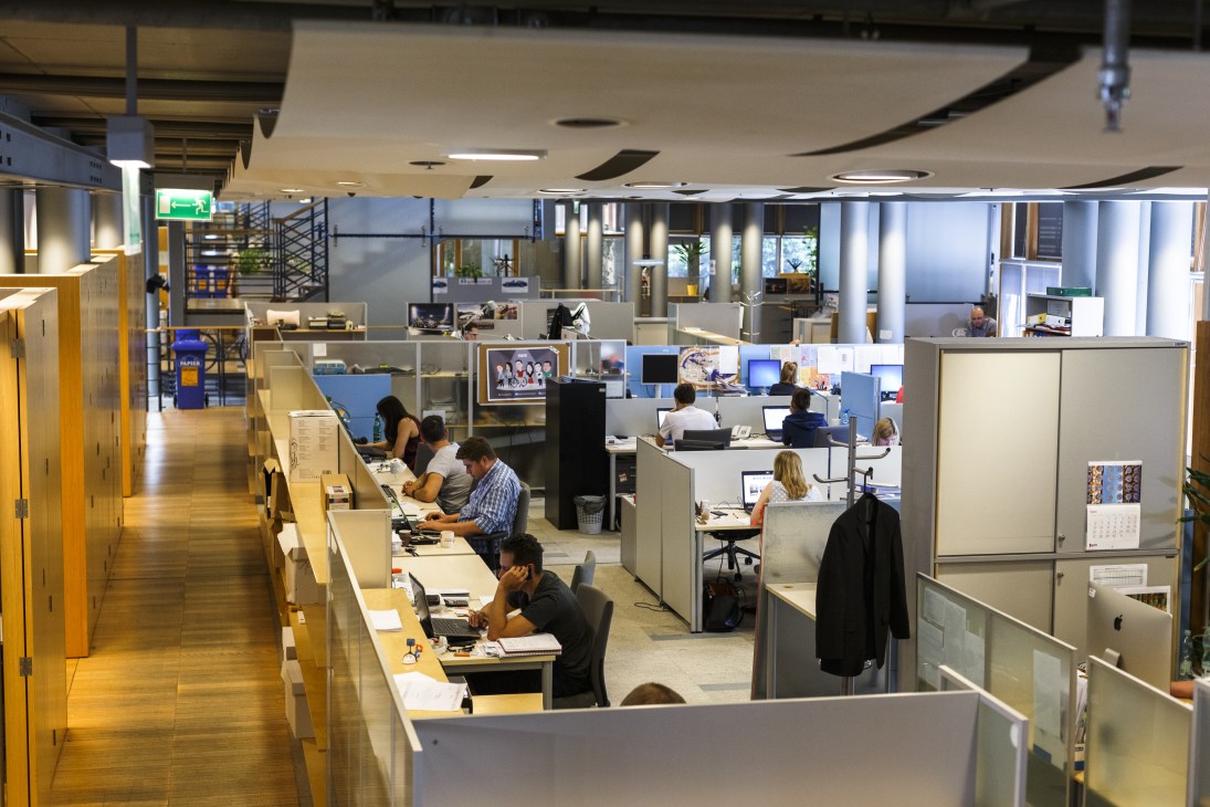 Open-plan editorial office with journalists working at desks separated by low partitions, yellow storage units, and natural lighting.
