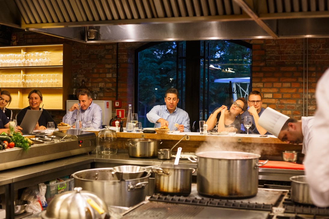 Diners seated at a bar counter surrounding an open kitchen at Warszawa Wschodnia restaurant.