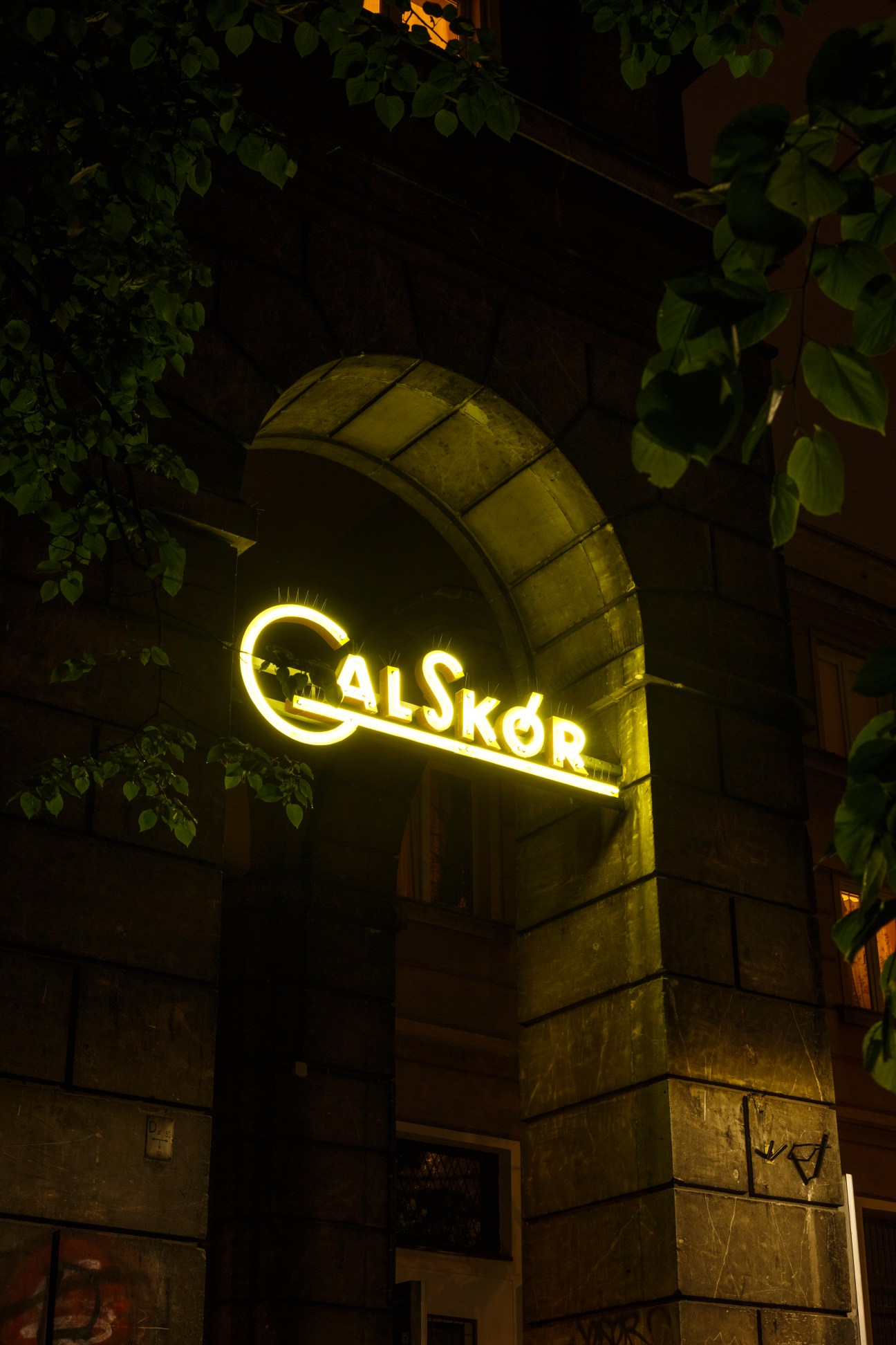 Illuminated yellow neon sign reading 'GALSKOR' on an arched storefront at night, surrounded by climbing ivy.