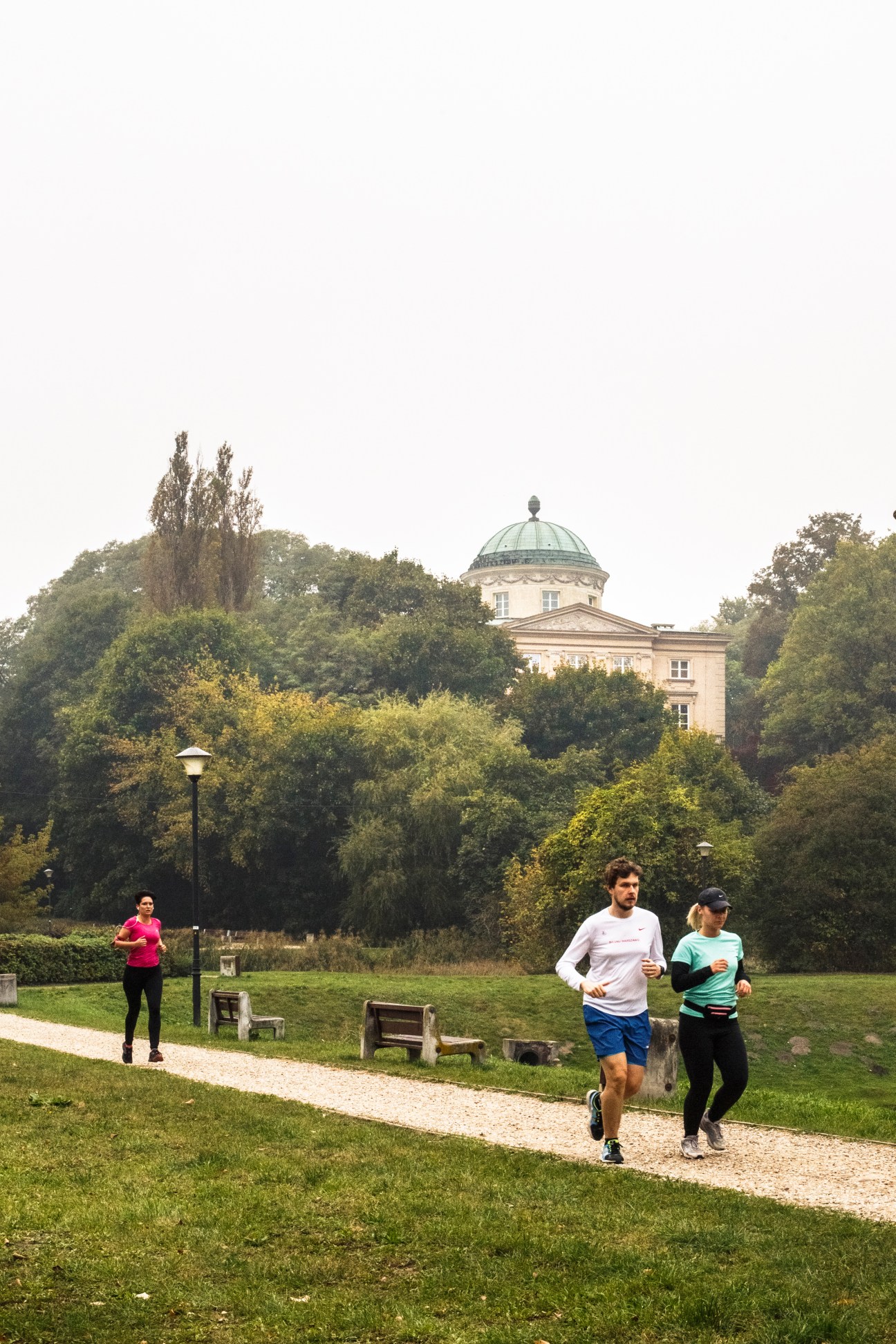 Joggers running on a tree-lined path in Arkadia Park with a domed classical building visible among green foliage.