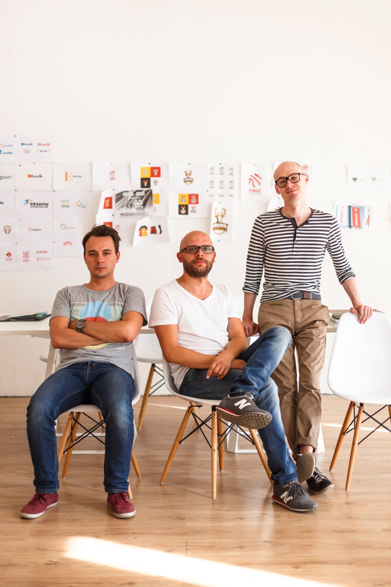 Three men pose in a minimalist office space with design work displayed on the wall behind them.