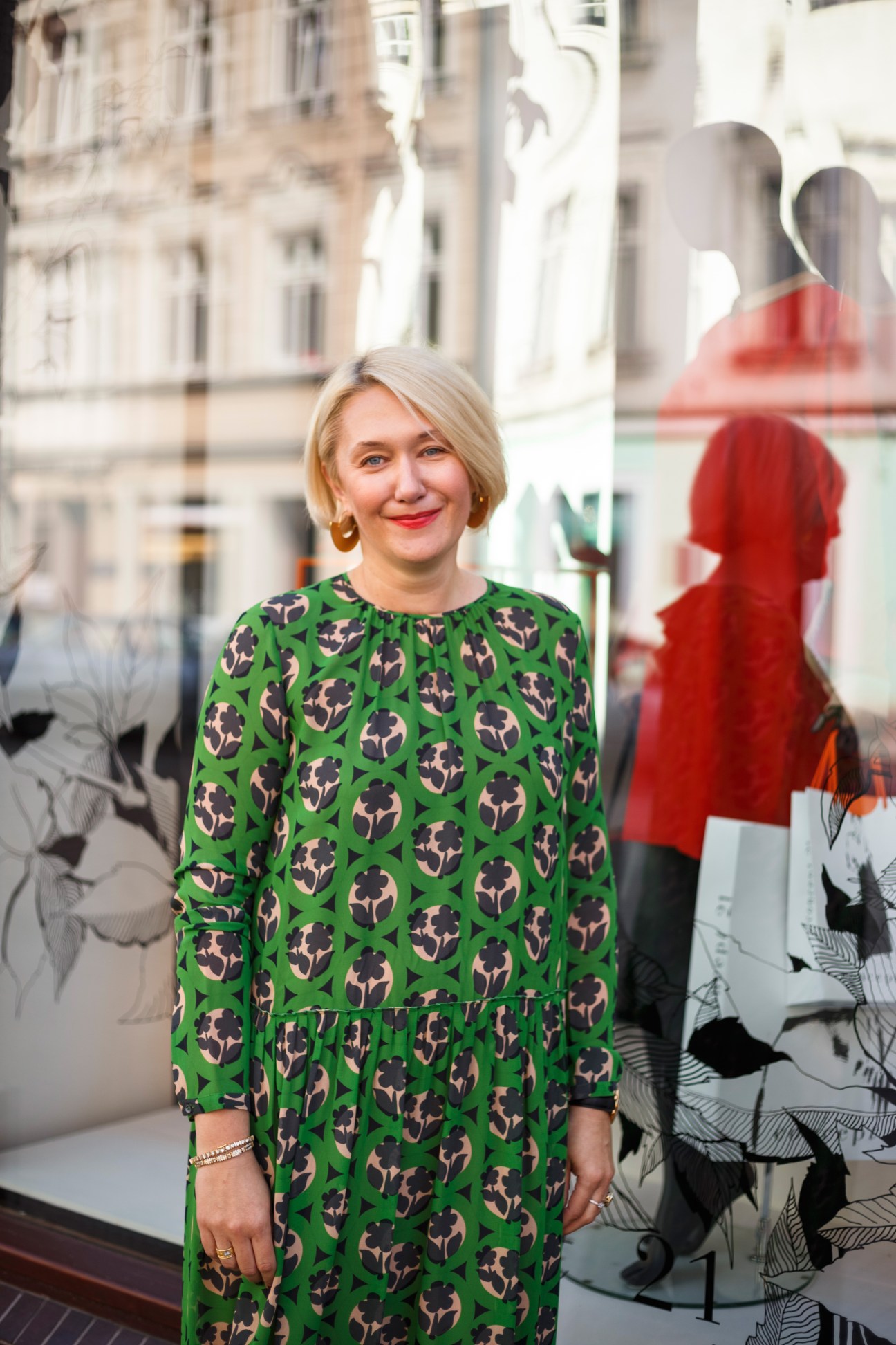 Woman in green patterned dress standing on city street with shop windows behind her.