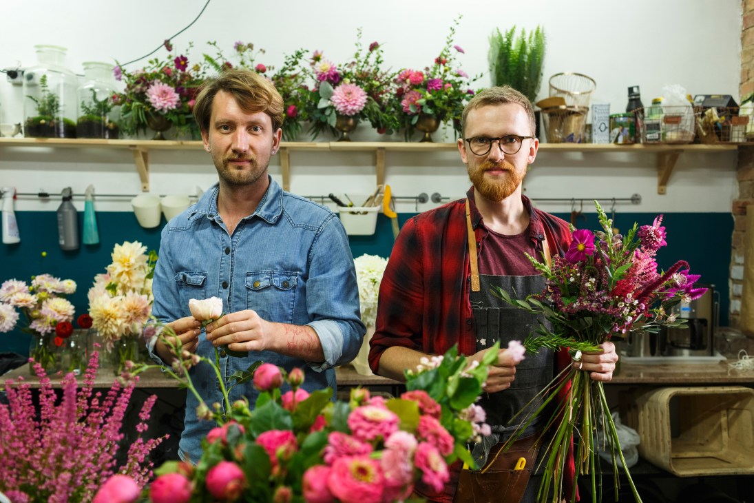 Two male florists arrange flowers in a shop with shelves of blooms and supplies behind them.
