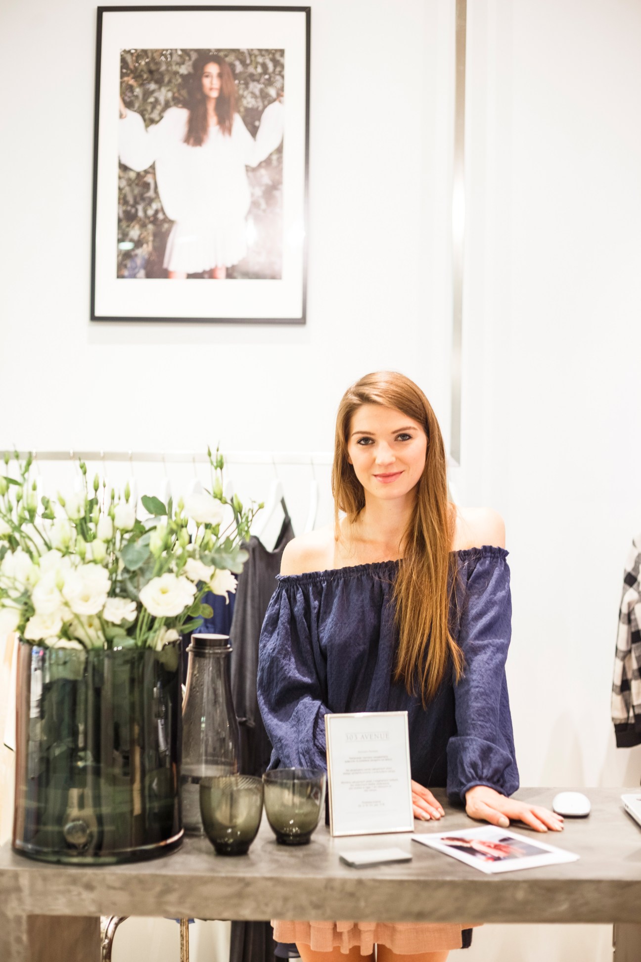 Young woman with long reddish-brown hair wearing a dark blue off-shoulder blouse stands at a counter in a minimalist retail space with flowers and fashion items displayed.