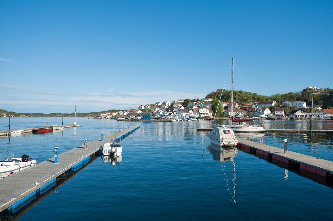Sailboats moored at a wooden dock in Kragerø's harbor with colorful waterfront houses on hillside beyond.