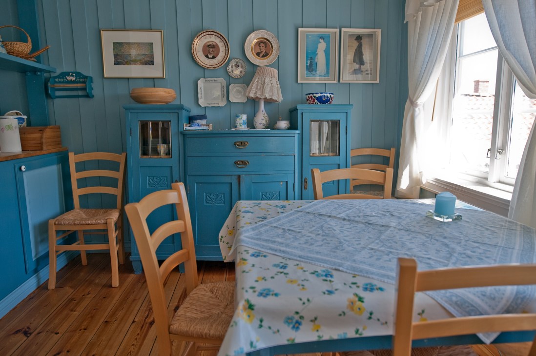 Bright turquoise bedroom with wooden furniture, framed artwork on walls, and windows overlooking coastal scenery.