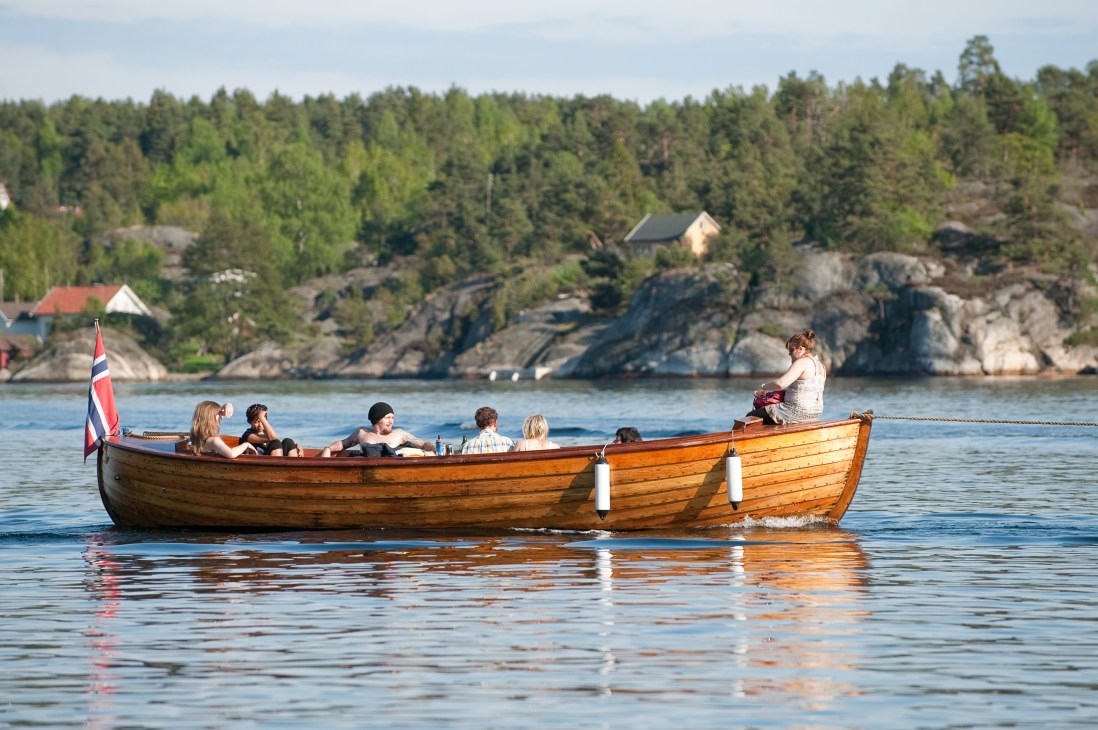 People in a wooden boat with Norwegian flag on a fjord with forested shoreline and houses in the background.