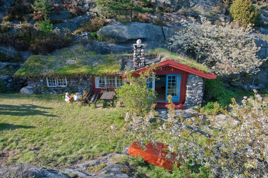 Red cabin with blue door nestled among boulders and flowering shrubs in a Norwegian coastal landscape.