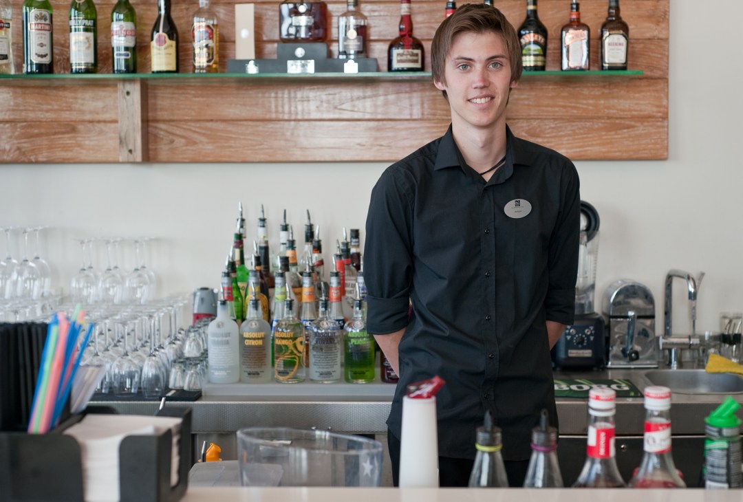 Bartender in black shirt standing behind a bar counter with bottles and glasses.