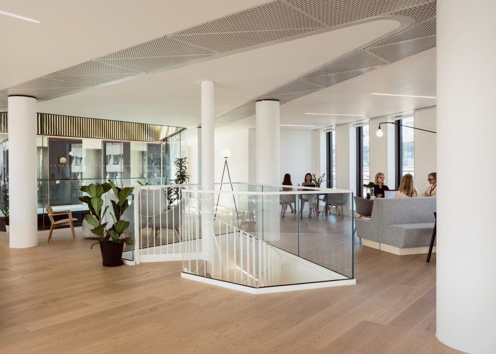 Modern open office space with natural light, white columns, potted plants, and employees working at desks behind glass partitions.