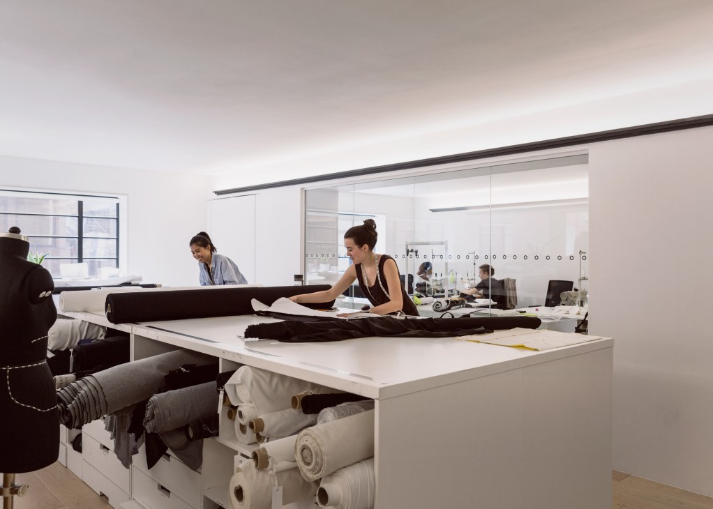 Pattern cutters working with fabric at desks in a bright, minimalist workspace with glass partitions.