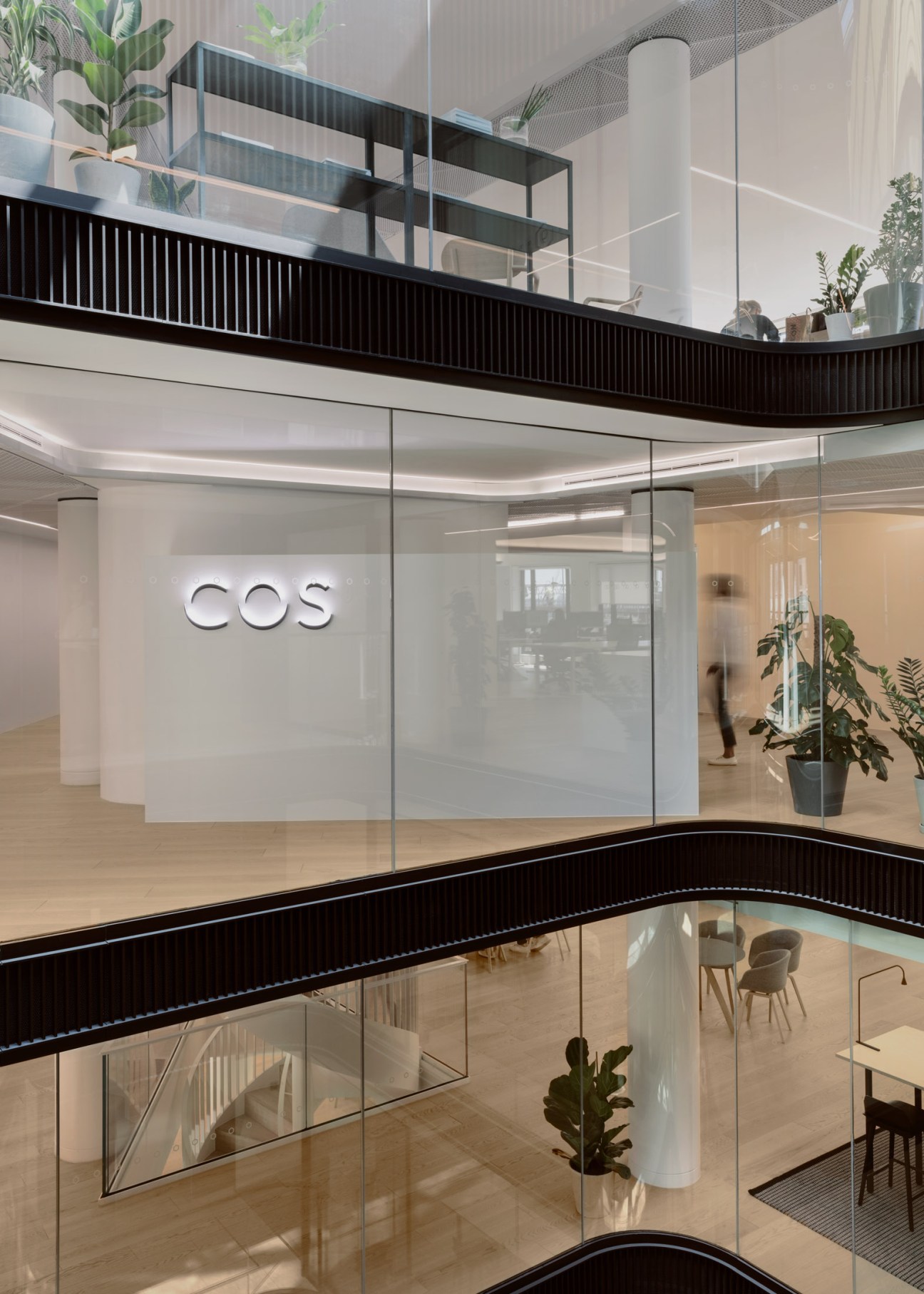Multi-level interior of Cos headquarters showing modern office space with glass railings, potted plants, and COS signage.