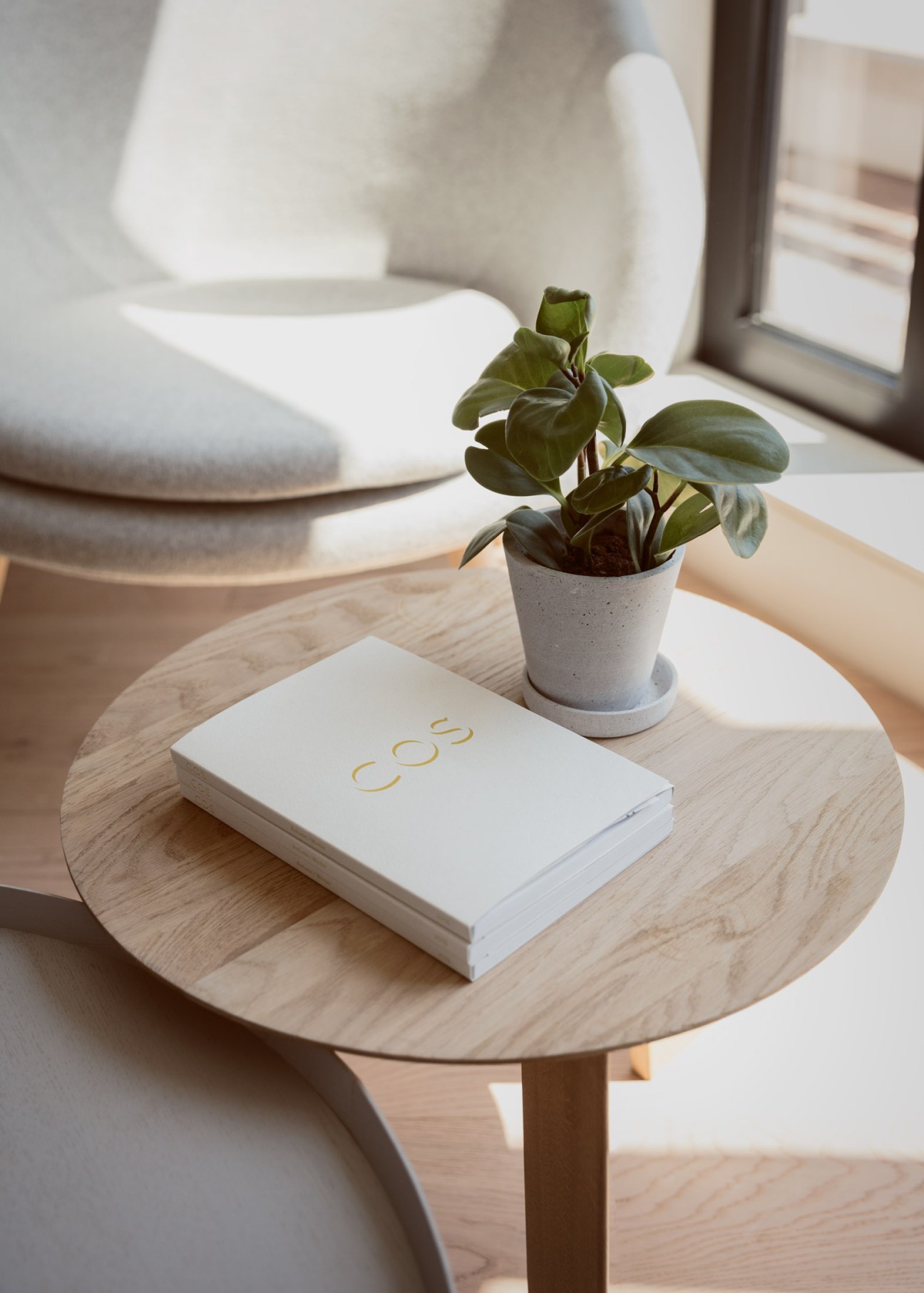 A minimalist wooden table displaying a white Cos notebook and potted plant beside a modern white chair.