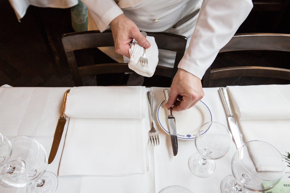 Server in white shirt placing napkin on place setting at formally dressed dining table.