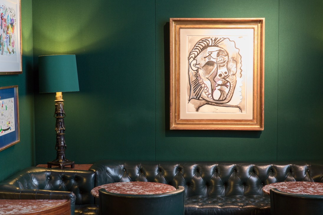 Framed abstract drawing by Picasso depicting a stylized head in brown ink on cream paper, hung on a deep green wall above a black leather tufted sofa.