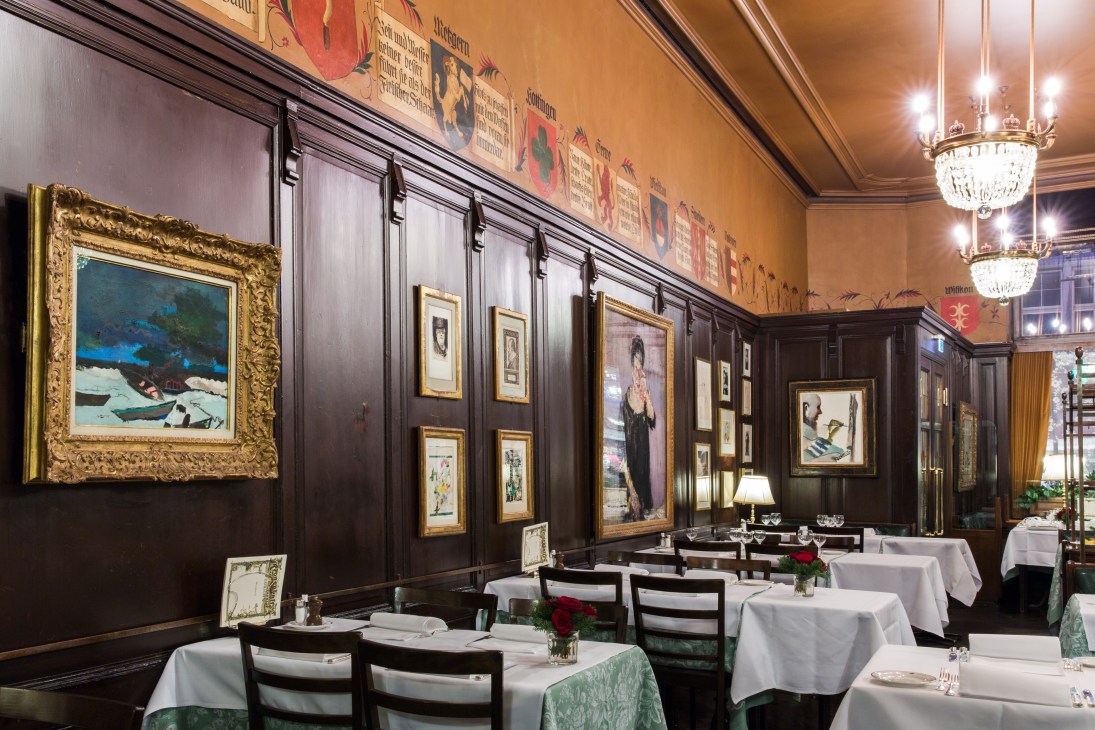 Elegant dining room at Kronenhalle with dark wood paneling, framed artworks on walls, white tablecloths, and crystal chandelier.