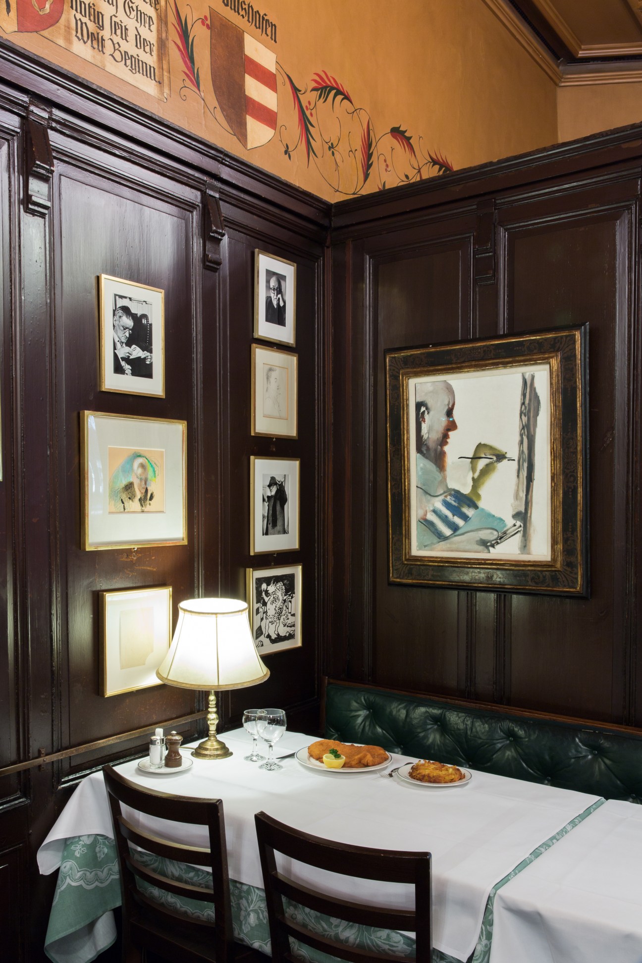 Dark wood-paneled dining room with framed artworks on walls, white tablecloth table set with lamp and food plates.
