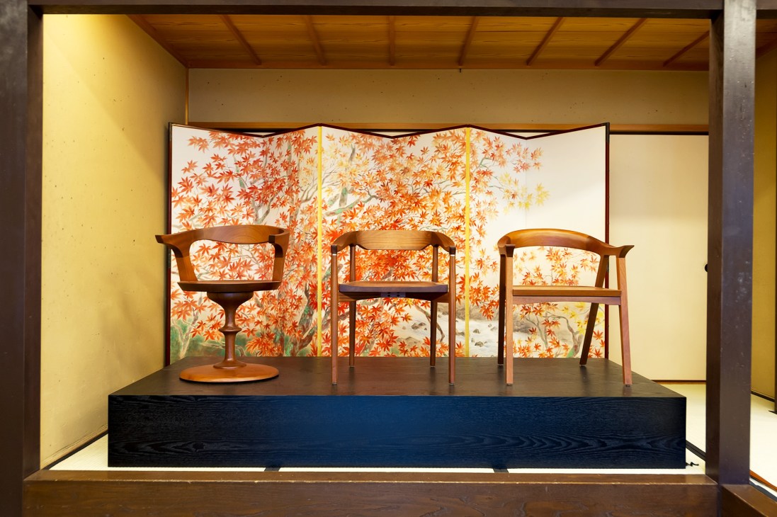 Stellar Works wooden chairs displayed on a black platform with a folding screen featuring orange maple leaf patterns in the background.