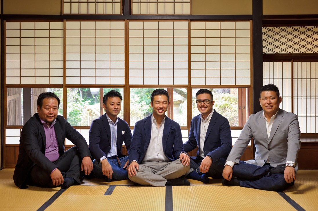 Five Japanese craftsmen in business attire sit cross-legged on a tatami mat in front of traditional shoji windows.