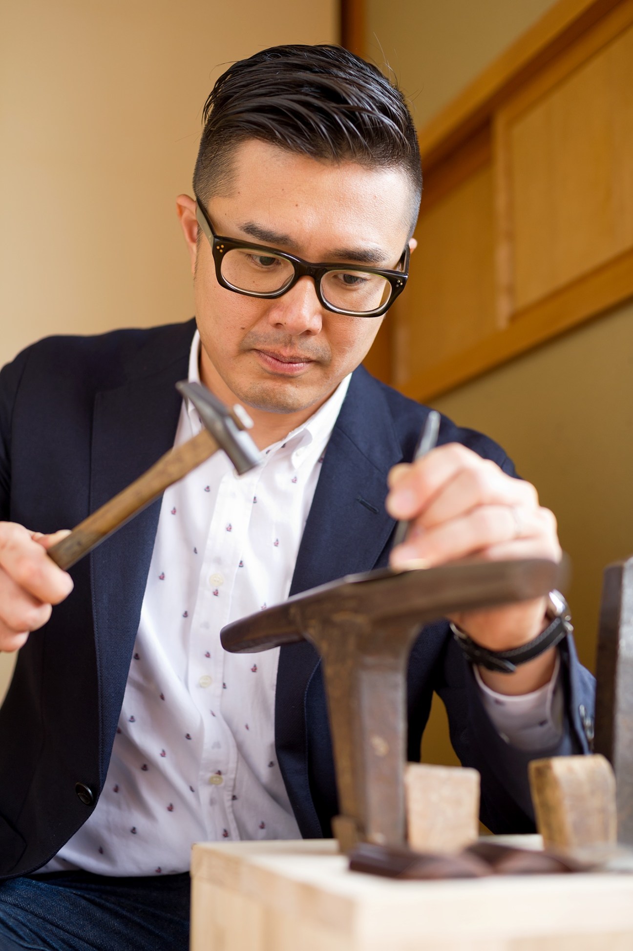 Takahiro Yagi, a craftsman wearing glasses and dark blazer, works with woodworking tools on wooden pieces.