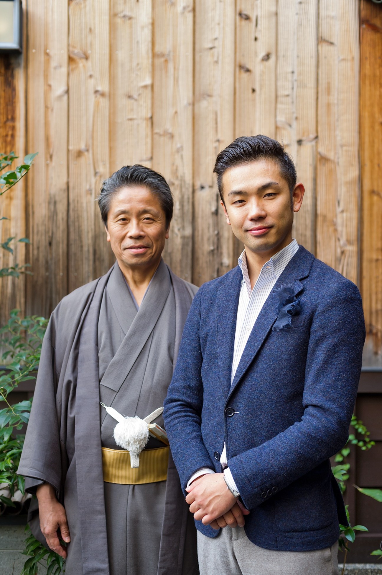 Masataka Hosoo (right) with his father, Masao Hosoo standing in front of a wooden wall.