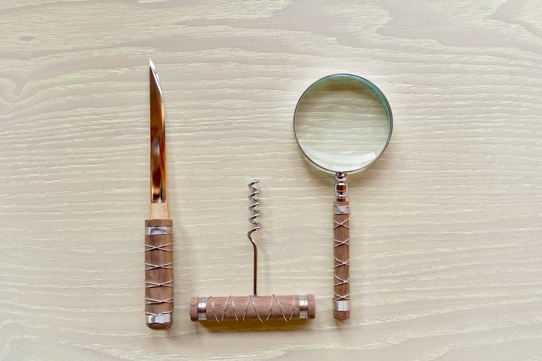 Three metal kitchen utensils with wooden handles wrapped in wire displayed on a light wooden surface: a knife, corkscrew, and magnifying glass.