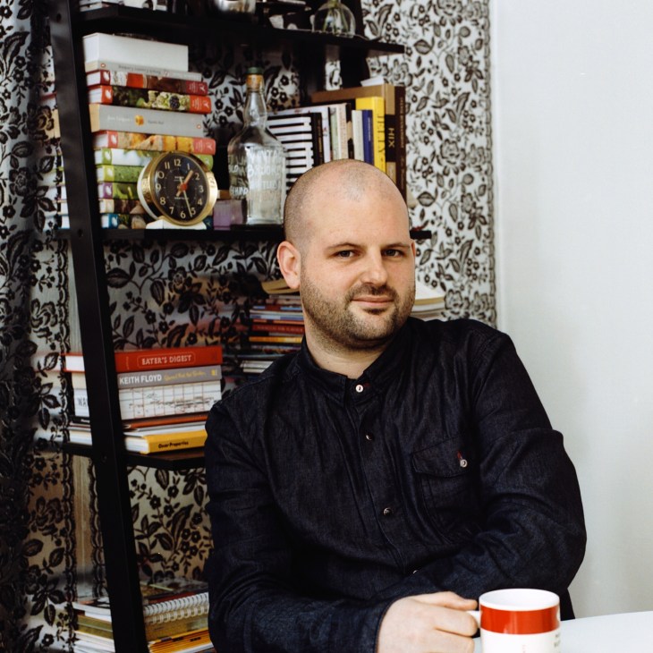 Man in dark denim shirt sits at table with patterned wallpaper and bookshelf behind him.