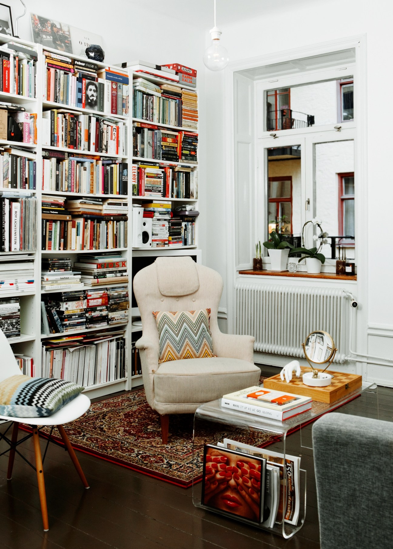 Modern living room with floor-to-ceiling white shelving filled with books, a cream armchair with patterned pillow, and large window overlooking Stockholm.