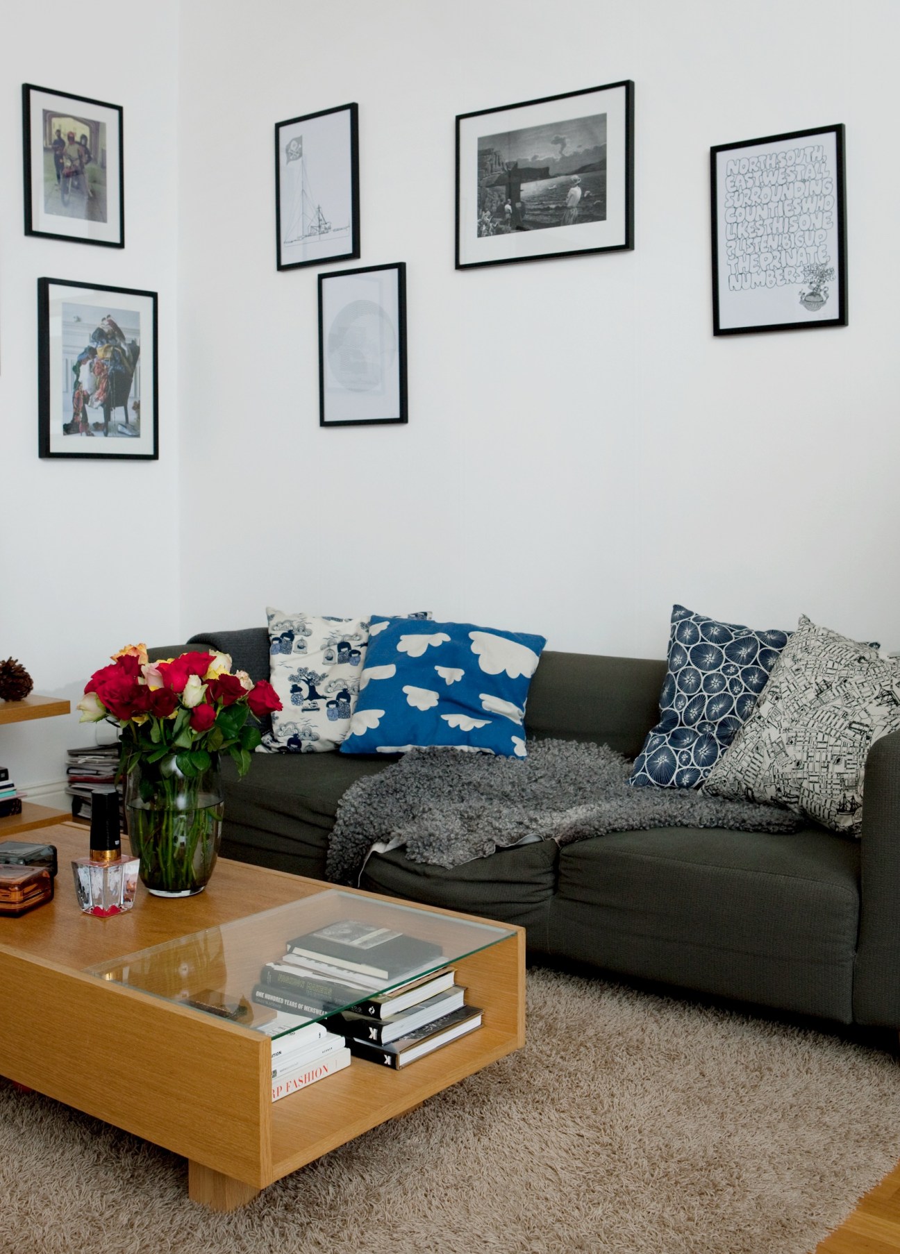 Modern living room with charcoal sofa, patterned cushions, wooden coffee table with books, fresh flowers, and framed artwork on white walls.