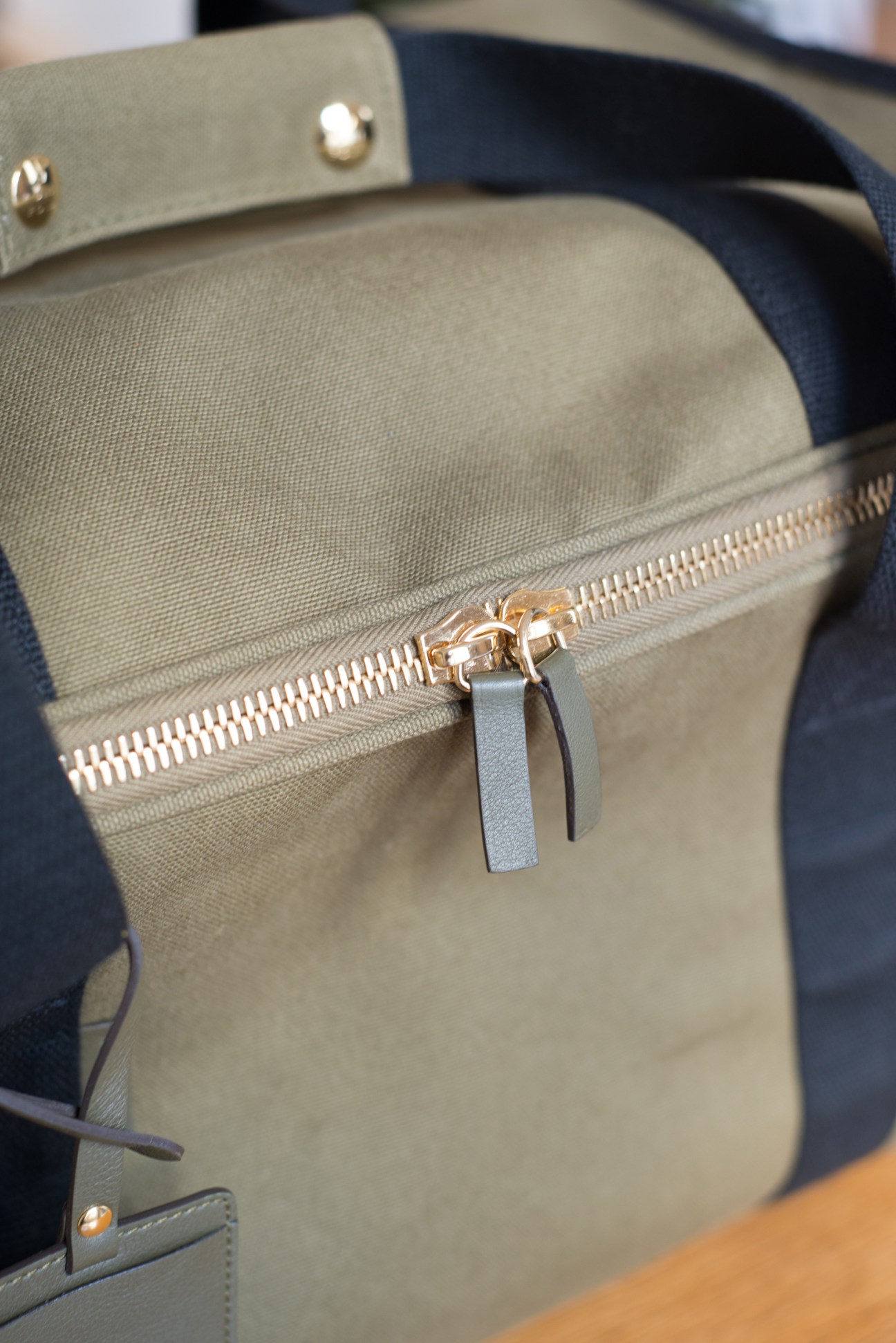 Close-up of a gold zip with fabric pulls on a beige canvas L/Uniform travel bag.