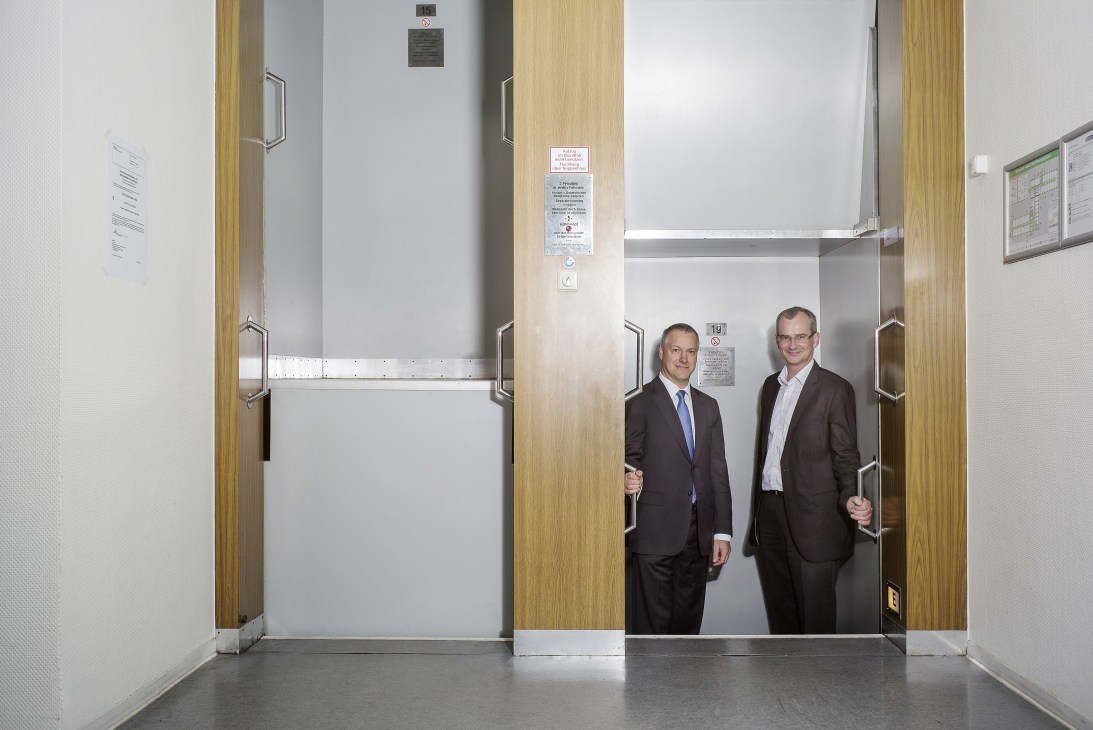 Two men in business suits stand in a paternoster lift with wooden and metal framing at ThyssenKrupp Elevator headquarters.