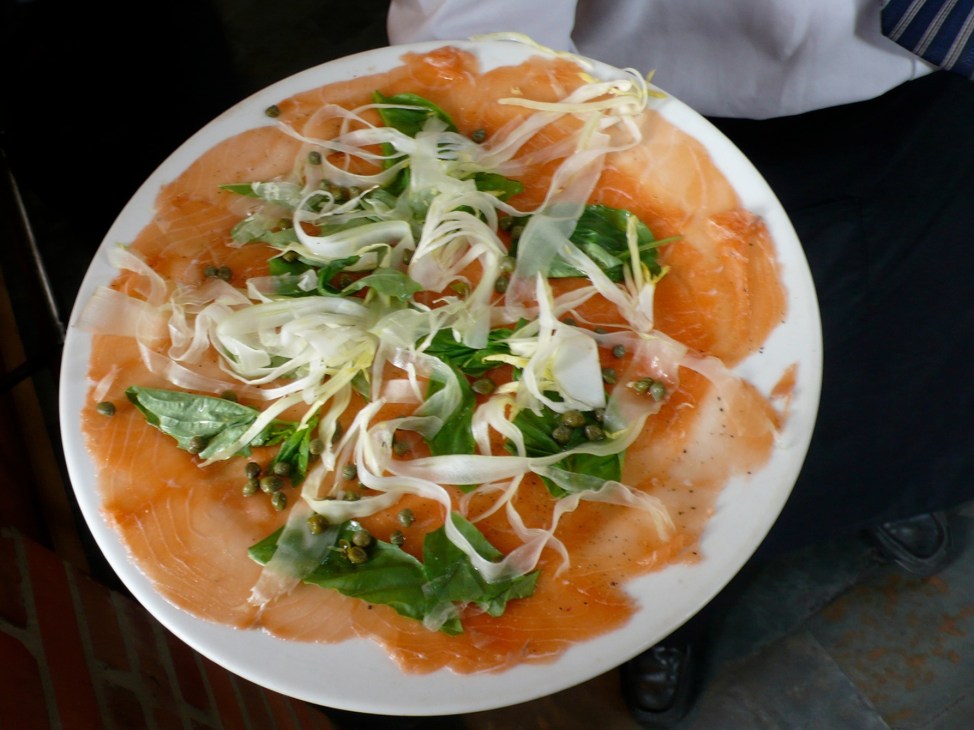Salmon carpaccio with sliced onions, capers, and fresh herbs on a white plate.