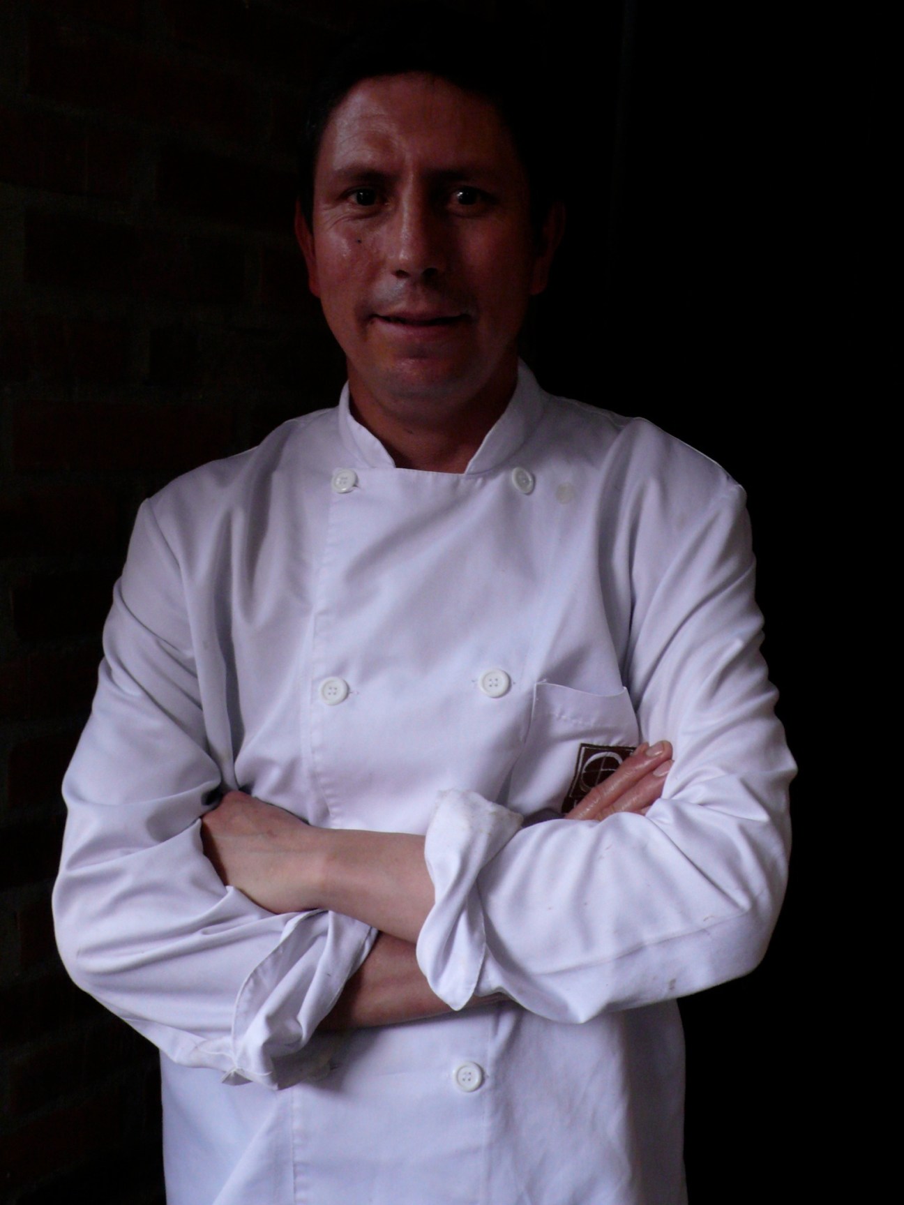 Chef Alberto Sanchez in white double-breasted chef's coat, arms crossed, against dark background.