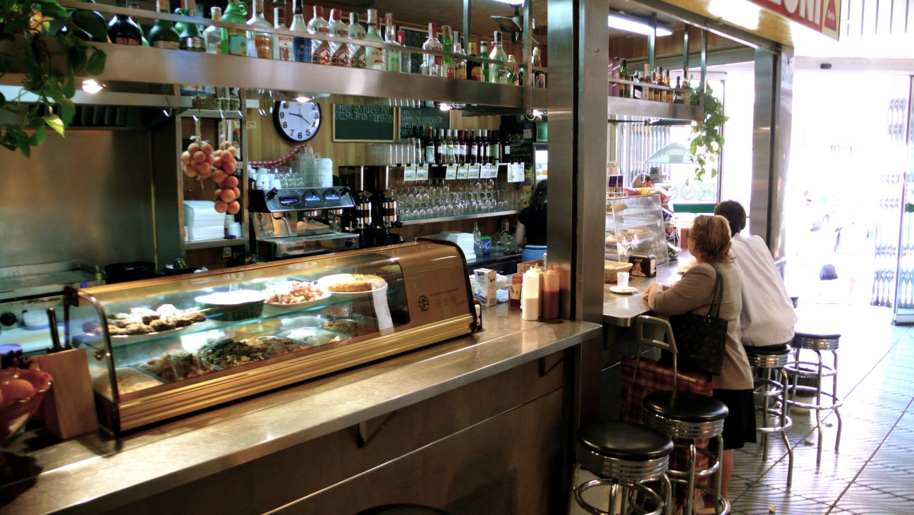 Interior of a café or deli counter with display cases, shelving, and customers seated at the bar.