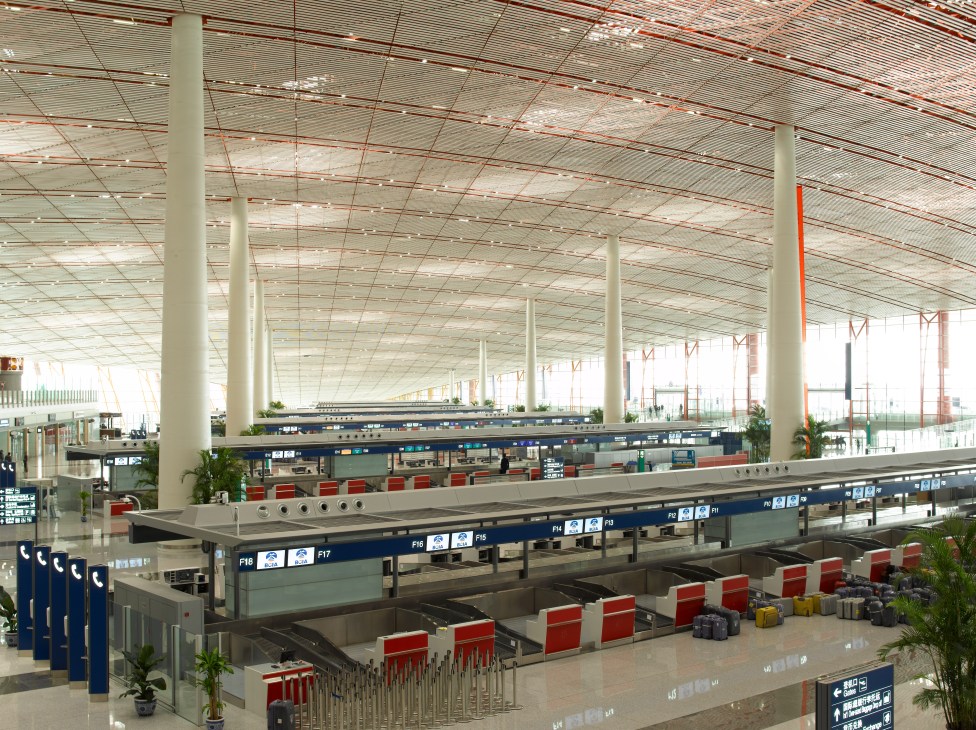 Interior of Terminal 3 at Beijing Capital Airport showing check-in counters, red seating, and a vast gridded ceiling with support columns.