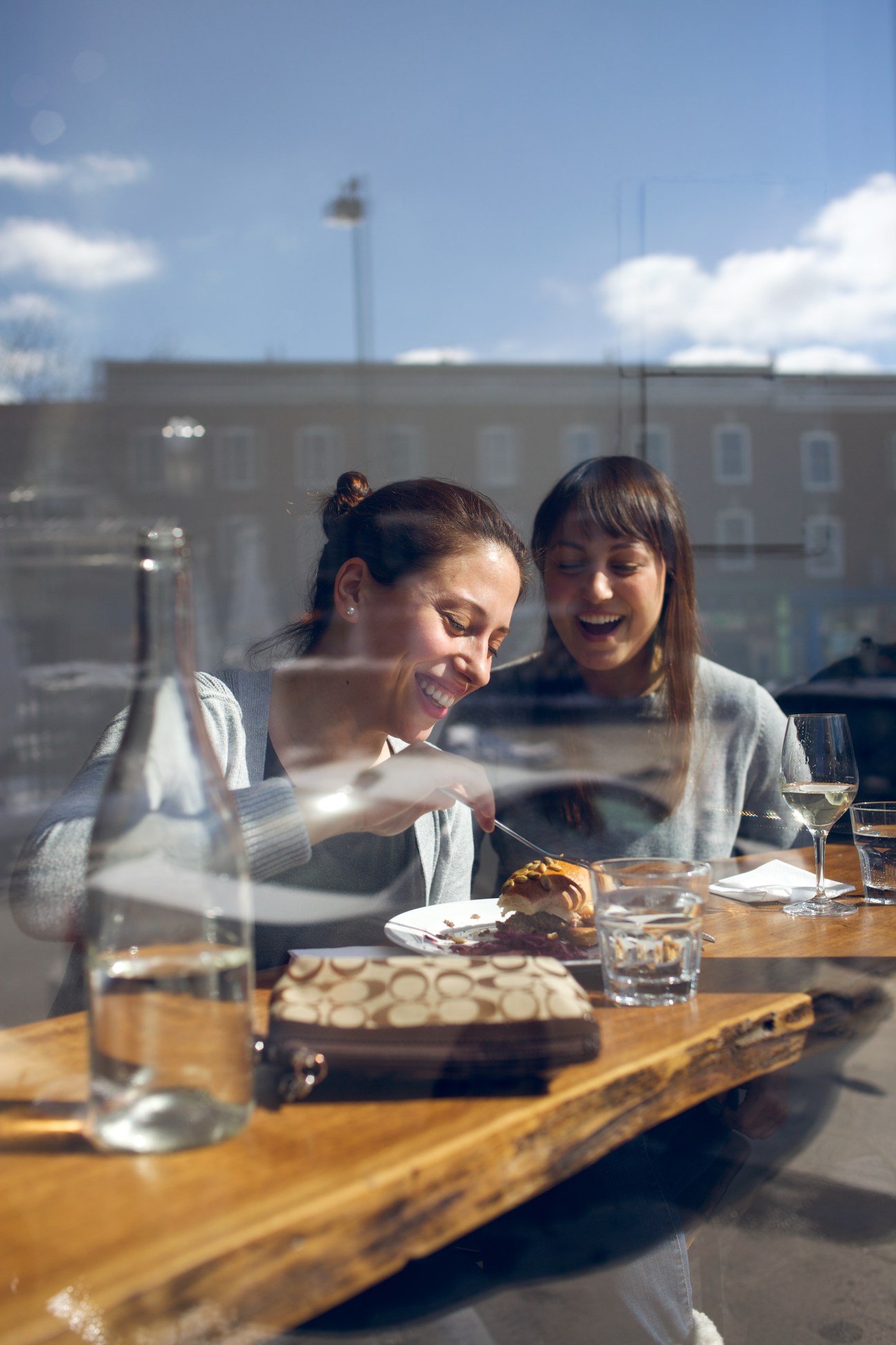 Two women dining at a wooden table with white wine and food, viewed through a window with city buildings behind.