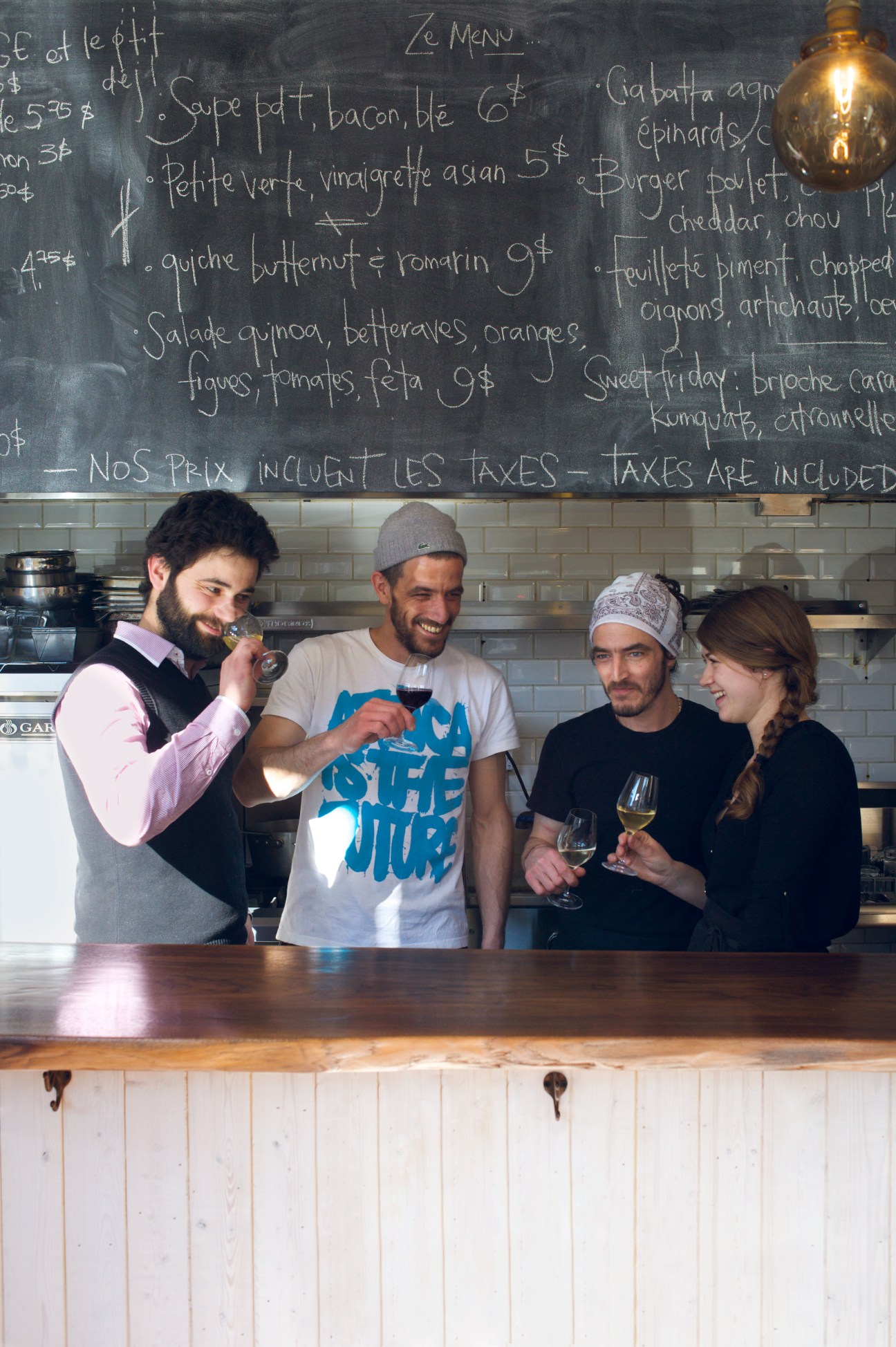 Four people smiling at a wooden counter in front of a chalkboard menu at La Famille restaurant in Montreal.