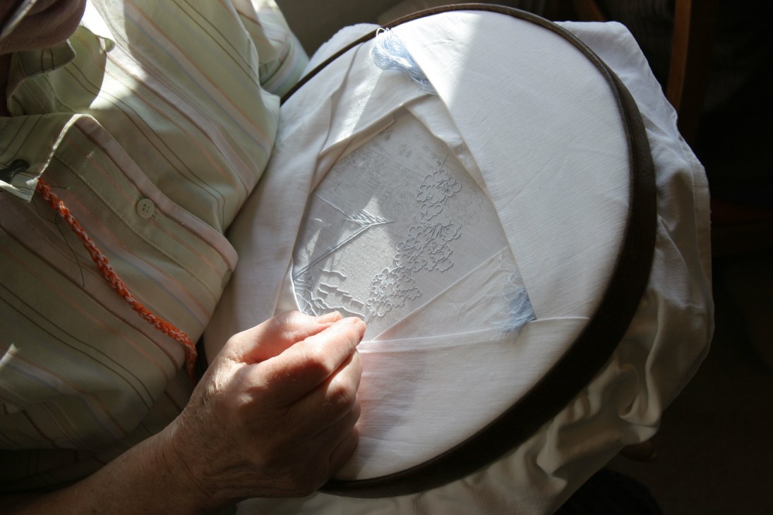 Close-up of an embroidered linen with detailed needlework held in a hoop by a seamstress's hands.